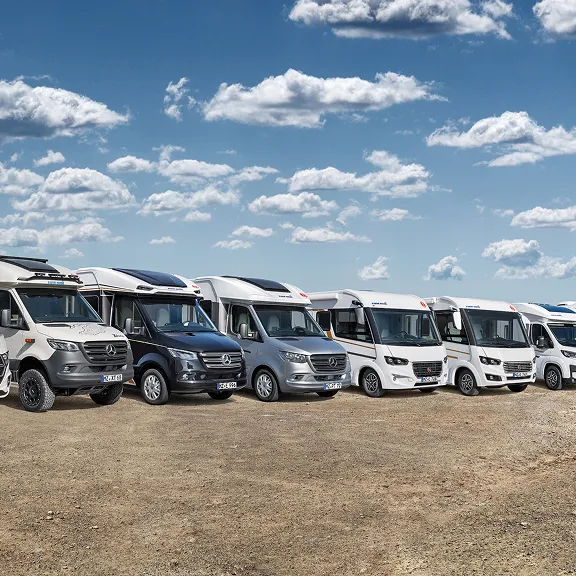 Reihe von sechs Wohnmobilen verschiedener Modelle und Größen, geparkt auf einem großen Asphaltplatz am Meer bei klarem Himmel.